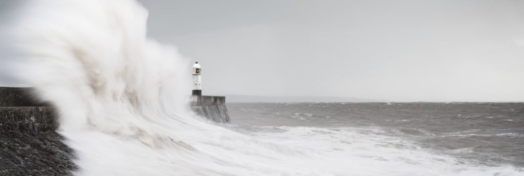 Waves crashing over the breakwater at Porthcawl, a small lighthouse in the distance