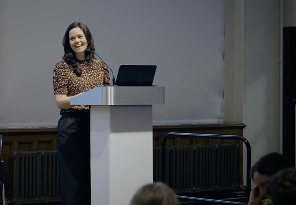 A woman stands at a lectern speaking from a stage.