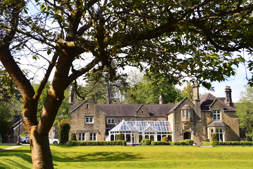 Large stone mansion with a glass conservatory, framed by a large tree, set on a manicured green lawn with trimmed hedges and parked cars visible in the distance.