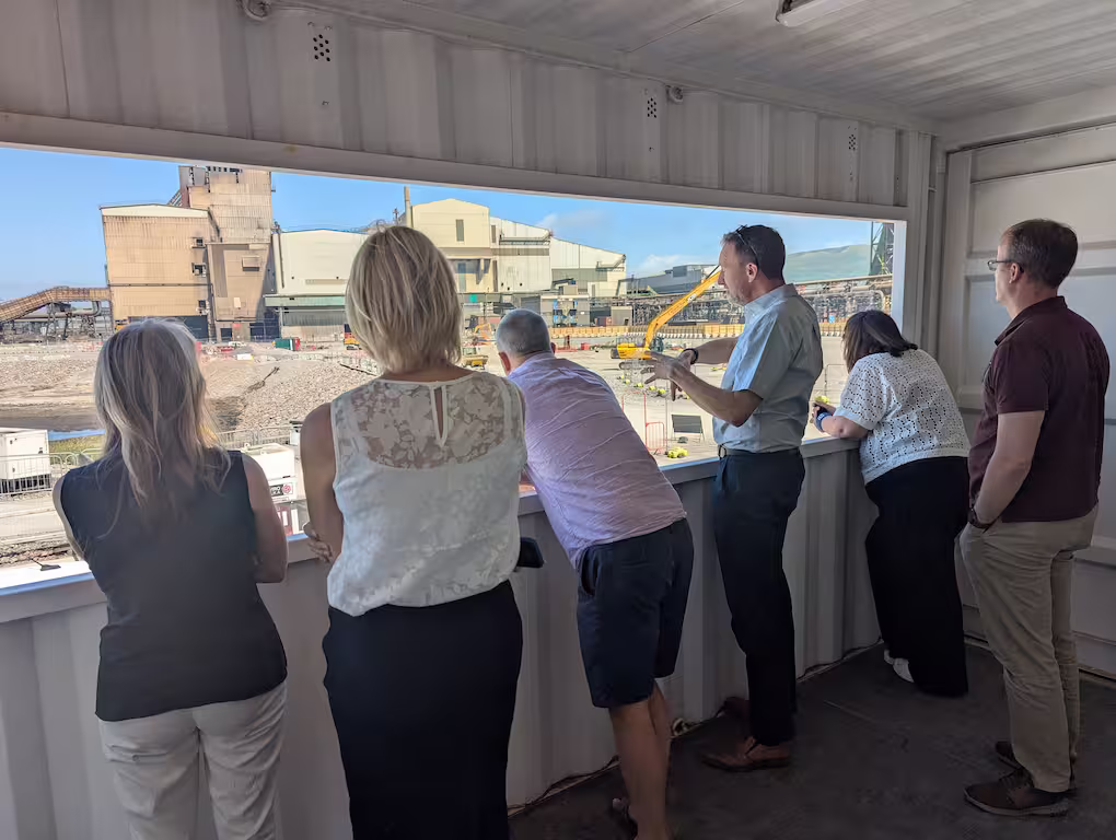 A group of people stands inside a room with a large window, looking out at an industrial complex with a large building, construction equipment, and a rocky landscape under a clear blue sky.