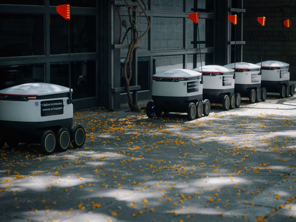 A line of autonomous delivery robots with red flags on top parked on a sidewalk covered in fallen yellow leaves in front of a building with a glass garage door.